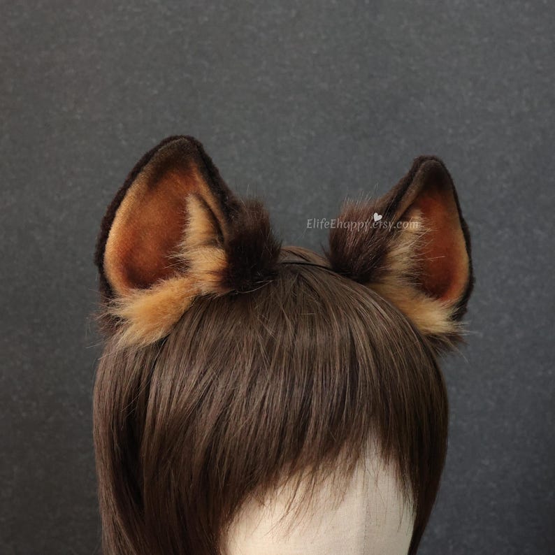 May include: A pair of brown and tan faux fur animal ears on a headband. The ears are detailed with darker brown tips and a lighter tan interior. The headband is worn on a person with brown hair.