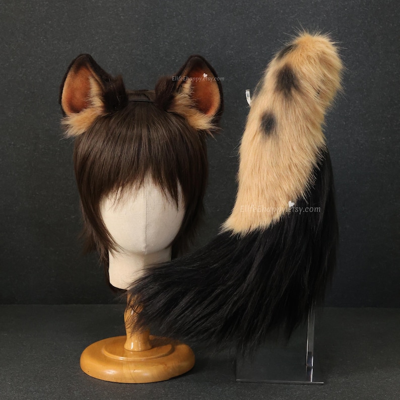 May include: A brown and tan faux fur cosplay set, including a pair of ears and a tail. The ears are brown with tan inner details. The tail is tan with black spots and a black base. The set is displayed on a mannequin head and a clear stand.