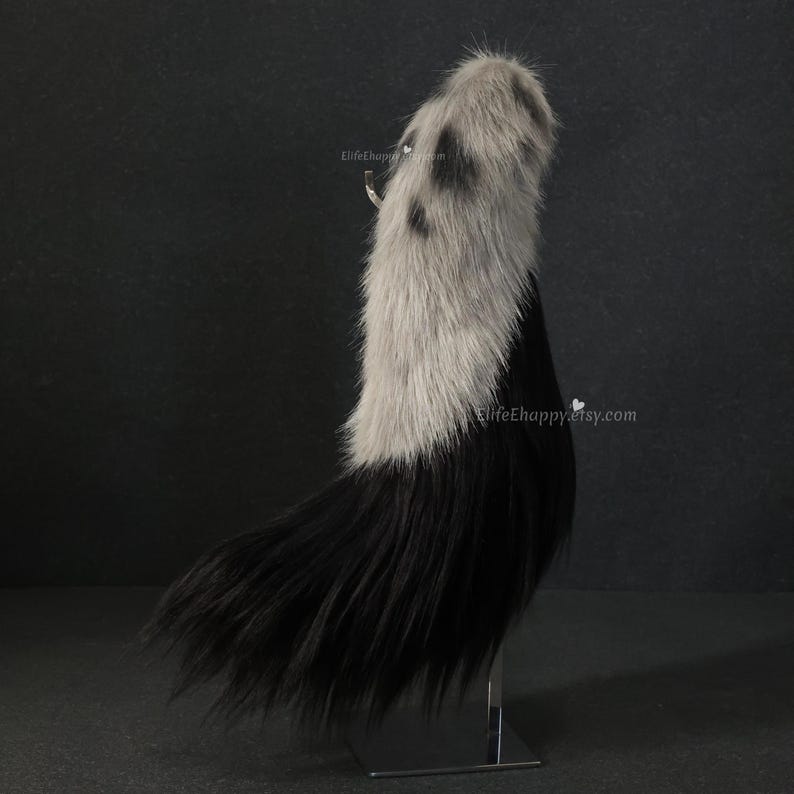 May include: A faux fur tail, primarily black with a gray and black spotted top section. The tail is displayed upright on a metal stand against a dark background. The Etsy shop name is visible.