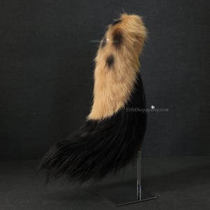 May include: A faux fur tail with a two-tone design. The top section is tan with black spots, transitioning to a black base. The tail is displayed on a silver stand against a dark background. The Etsy shop name is visible.