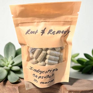 Può includere: Un sacchetto di carta marrone con una finestra trasparente contenente capsule. Il sacchetto è etichettato "Root & Remedy" e "Indigestion Capsules 50 count."