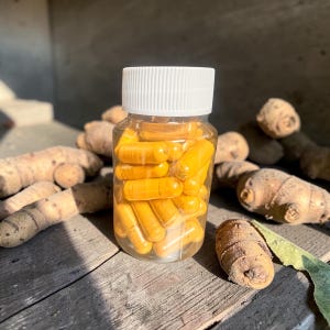 Może przedstawiać: Przezroczysta plastikowa butelka wypełniona jasnożółtymi kapsułkami, zwieńczona białą pokrywką. Butelka stoi na zwietrzałej drewnianej powierzchni, otoczona kilkoma kawałkami surowego korzenia kurkumy.