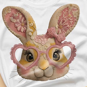 Puede incluir: Camiseta blanca con una ilustración detallada de una cara de conejo con gafas en forma de corazón y un lazo rosa. El conejo tiene ojos grandes y está decorado con motivos florales rosas y amarillos.