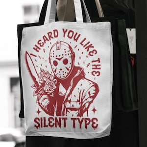 Puede incluir: Bolso tote blanco con un gráfico rojo de una figura enmascarada sosteniendo un cuchillo y rosas. El texto en el bolso dice "I HEARD YOU LIKE THE SILENT TYPE". Tiene asas a rayas blancas y negras.