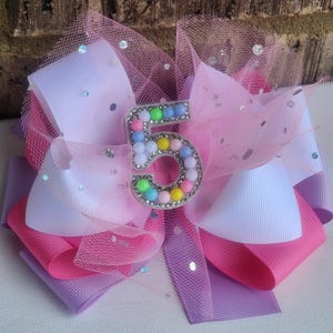 May include: A large, layered hair bow in shades of pink, white, and lavender, accented with a sparkly tulle overlay. The bow features a rhinestone-studded number "5" in the center, with colorful bead accents. Scattered sequins add extra sparkle.