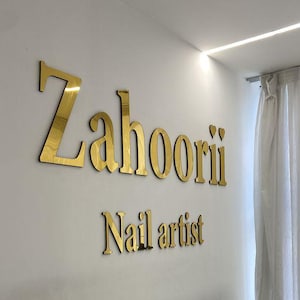Op de afbeelding: Interieur van een nagelsalon met gouden letters op een witte muur met de tekst "Zahoorii Nail artist". Een tafel met glazen blad met nagelverzorgingsapparatuur staat op de voorgrond. De kamer heeft een moderne, strakke uitstraling met heldere bovenverlichting.