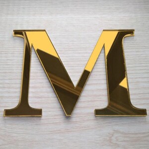 May include: A gold-colored letter "M" made of reflective material. The letter is displayed on a light-colored wooden surface. The capital letter has a classic serif font.