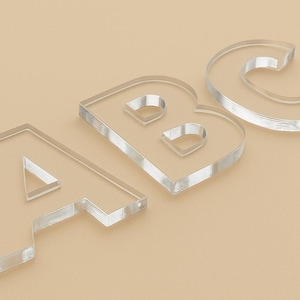 Puede incluir: Letras A, B y C de acrílico transparente sobre un fondo beige. Las letras tienen un aspecto esmerilado con bordes biselados, creando un efecto tridimensional. Ideales para manualidades o proyectos educativos.