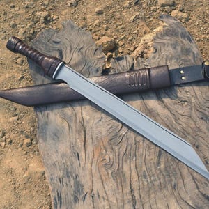 May include: A silver-bladed sword with a brown leather-wrapped handle and a matching sheath. The sword is laid on a weathered wooden surface. The sheath has a belt loop and decorative stitching.
