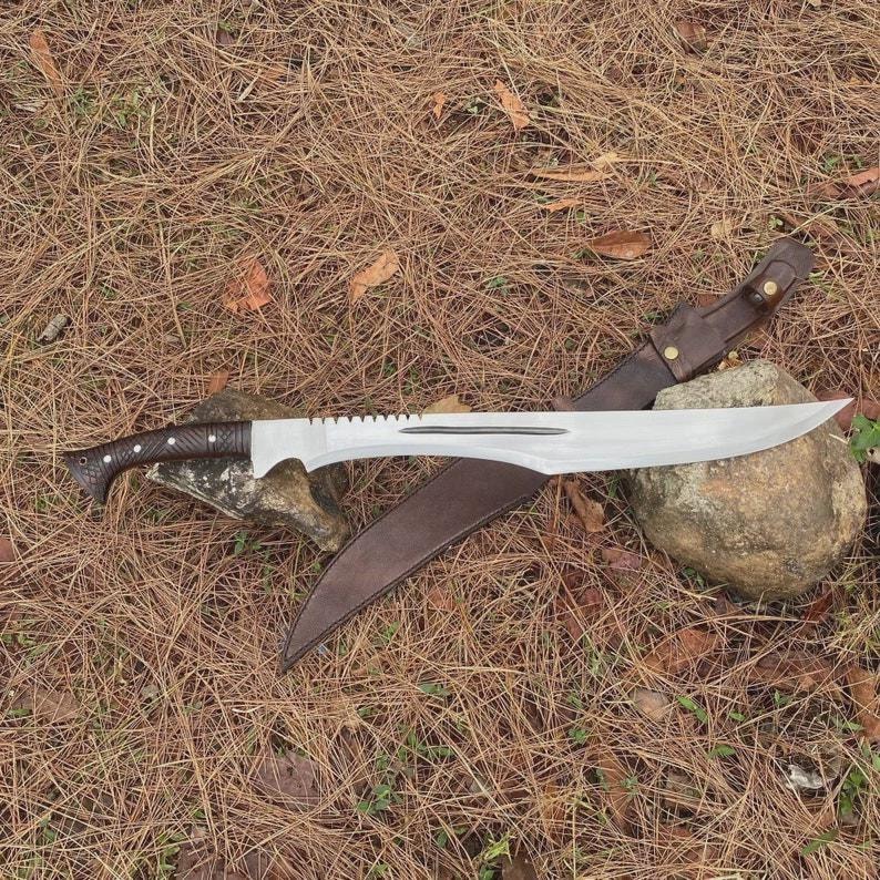 Puede incluir: Una gran machete plateado con un mango marr&oacute;n oscuro y un borde dentado. La hoja curva descansa sobre una funda de cuero marr&oacute;n. El machete se exhibe sobre un lecho de agujas de pino.