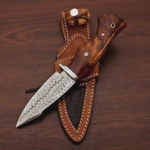 Puede incluir: Un cuchillo artesanal con una hoja estampada y un mango de madera, en una funda de cuero marr&oacute;n. La funda est&aacute; cosida con hilo blanco y tiene un cierre de bot&oacute;n a presi&oacute;n. La hoja del cuchillo tiene un patr&oacute;n ondulado &uacute;nico.