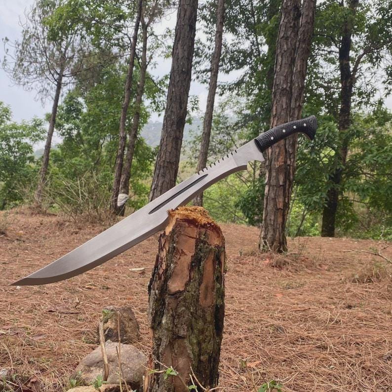 Puede incluir: Una gran machete plateado con un mango negro descansa sobre un toc&oacute;n de &aacute;rbol. La hoja tiene un borde dentado cerca del mango y una punta curva. El fondo presenta un bosque con &aacute;rboles altos y un entorno natural.