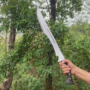 Puede incluir: Una gran machete plateado con una hoja curva y un borde dentado. El mango es negro y marr&oacute;n con un dise&ntilde;o texturizado. El machete se sostiene contra un tel&oacute;n de fondo de follaje y &aacute;rboles verdes.