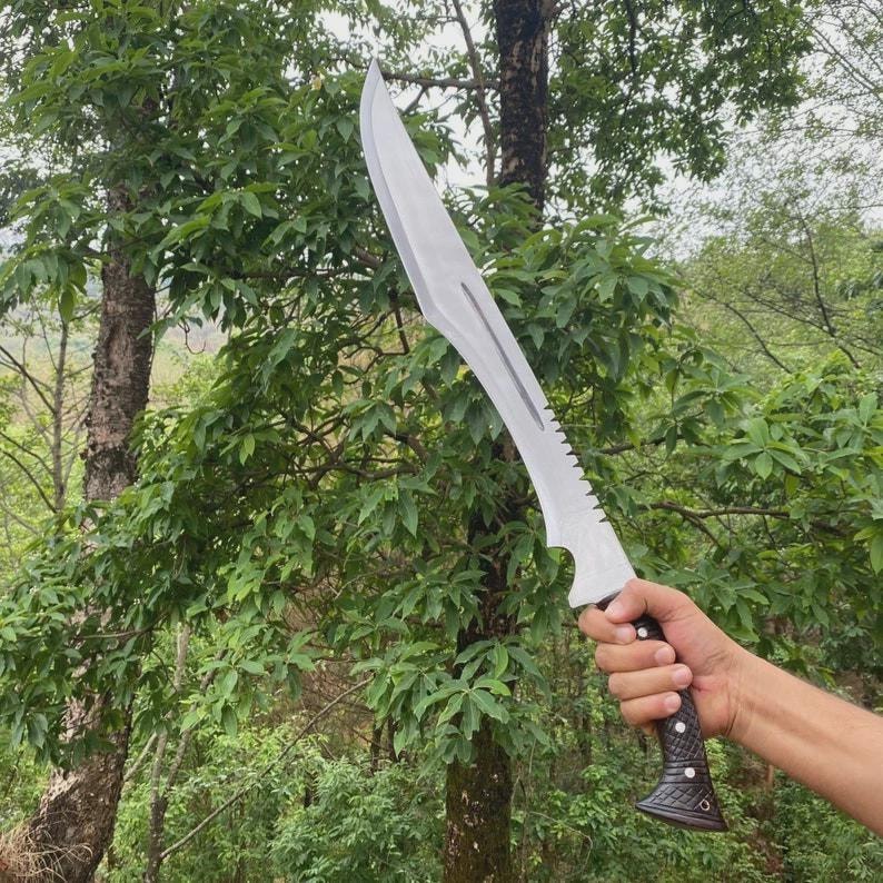 Puede incluir: Una gran machete plateado con una hoja curva y un borde dentado. El mango es negro y marr&oacute;n con un dise&ntilde;o texturizado. El machete se sostiene contra un tel&oacute;n de fondo de follaje y &aacute;rboles verdes.