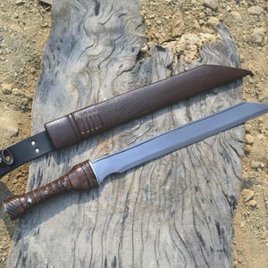 May include: A handmade sword with a brown leather-wrapped handle and a silver blade, resting on a weathered wooden surface. The sword is accompanied by a brown leather sheath and a black leather belt.