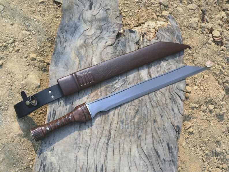 May include: A handmade sword with a brown leather-wrapped handle and a silver blade, resting on a weathered wooden surface. The sword is accompanied by a brown leather sheath and a black leather belt.