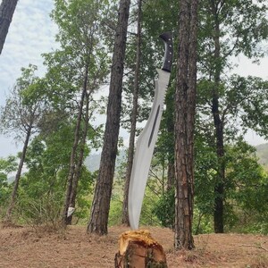 Puede incluir: Una gran machete curvada con un borde dentado y un mango oscuro y texturizado est&aacute; incrustada en un toc&oacute;n. La hoja es plateada, reflejando la luz. La escena est&aacute; ambientada en un bosque con &aacute;rboles altos y un cielo nublado.