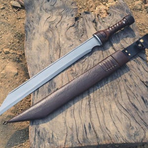 May include: A silver-bladed sword with a brown leather-wrapped handle and a matching sheath. The sword is lying on a weathered wooden surface, showcasing its sharp design and craftsmanship. The sheath has a leather strap with a buckle.