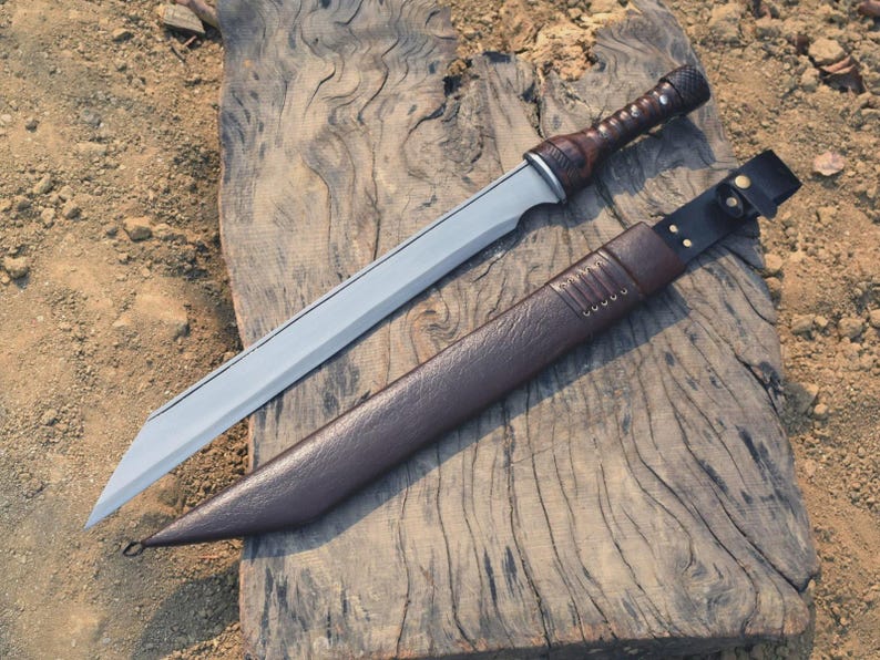 May include: A silver-bladed sword with a brown leather-wrapped handle and a matching sheath. The sword is lying on a weathered wooden surface, showcasing its sharp design and craftsmanship. The sheath has a leather strap with a buckle.