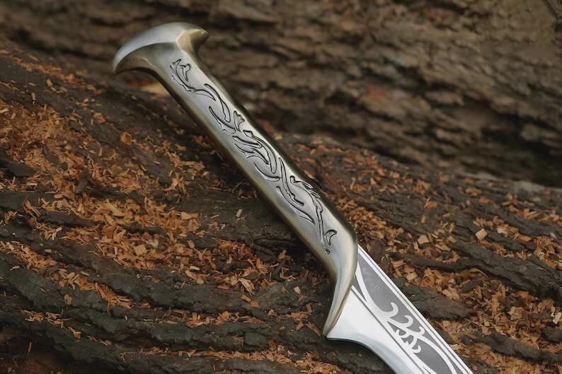 May include: Close-up of a silver knife with an ornate handle and blade. The handle features a curved design with etched floral patterns. The blade has a decorative pattern near the tip. The knife rests on a textured, brown wooden surface.