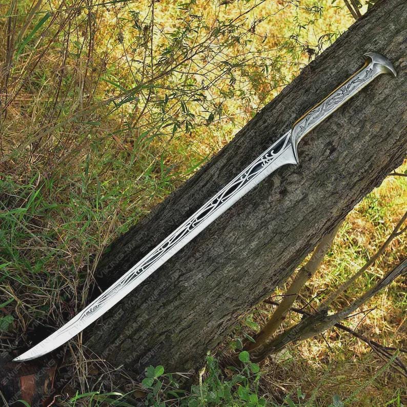 May include: A silver fantasy sword with an ornate handle and blade design. The blade has a curved edge and decorative cutouts. The sword is resting on a tree trunk, with greenery in the background.