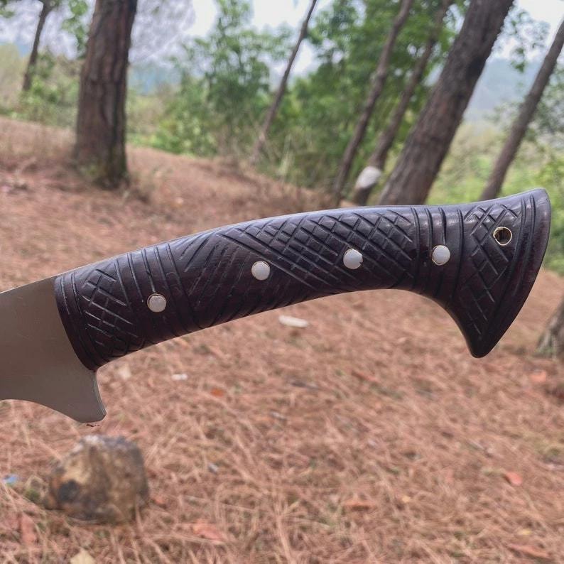 Puede incluir: Primer plano de un cuchillo con un mango marr&oacute;n oscuro texturizado. El mango presenta un patr&oacute;n tallado y tres remaches plateados. La hoja es plateada y parcialmente visible. El cuchillo est&aacute; sobre un fondo borroso al aire libre.