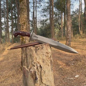 May include: A large, silver-colored sword with a brown handle and a leather sheath rests on a tree stump. The sword has a wide blade and a crossguard. The handle is intricately carved. The sheath is brown and has a loop at the end.