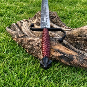 May include: A sword with a long, dark, textured blade featuring a serrated edge. The handle is wrapped in a deep red, spiraled material, complemented by a black metal crossguard and a pointed, multi-faceted black pommel at the base.