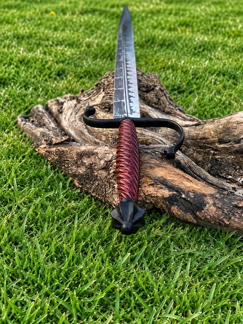 May include: A sword with a long, dark, textured blade featuring a serrated edge. The handle is wrapped in a deep red, spiraled material, complemented by a black metal crossguard and a pointed, multi-faceted black pommel at the base.