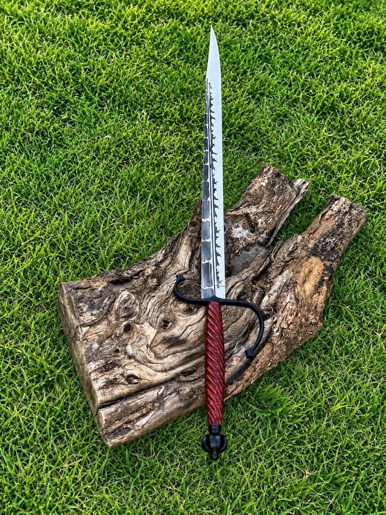 May include: A long, pointed sword with a silver-colored blade featuring a dark, textured spine. The handle is a deep red, twisted design with a black metal crossguard and a rounded black pommel at the base of the grip, creating a sharp, slender profile.
