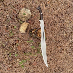 Puede incluir: Una gran hoja plateada con un borde dentado y un mango oscuro y texturizado. La hoja tiene un dise&ntilde;o curvo y una punta larga y afilada. El mango es negro y marr&oacute;n. El cuchillo est&aacute; sobre un lecho de agujas de pino marrones.