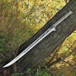 May include: A silver fantasy sword with an ornate handle and blade design. The blade has a curved edge and decorative cutouts. The sword is resting on a tree trunk, with greenery in the background.