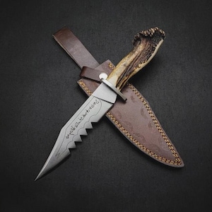 Puede incluir: Un cuchillo de caza con una hoja dentada y un mango de asta. El cuchillo está en una funda de cuero marrón con costuras decorativas. La hoja tiene texto grabado. El cuchillo está sobre un fondo oscuro.