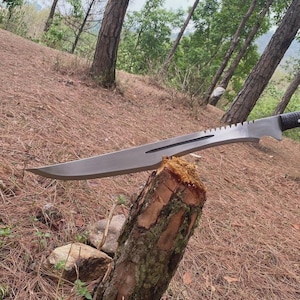 Puede incluir: Una gran machete plateado con lomo dentado y mango negro descansa sobre un toc&oacute;n. La hoja es larga y curvada, con una punta afilada. El fondo presenta un entorno forestal con &aacute;rboles y follaje marr&oacute;n.