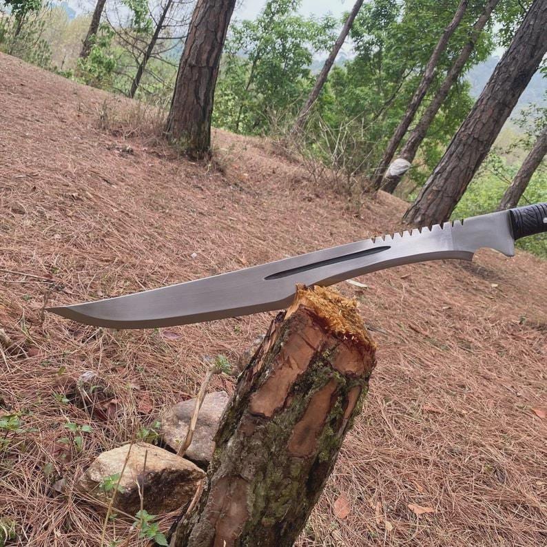Puede incluir: Una gran machete plateado con lomo dentado y mango negro descansa sobre un toc&oacute;n. La hoja es larga y curvada, con una punta afilada. El fondo presenta un entorno forestal con &aacute;rboles y follaje marr&oacute;n.