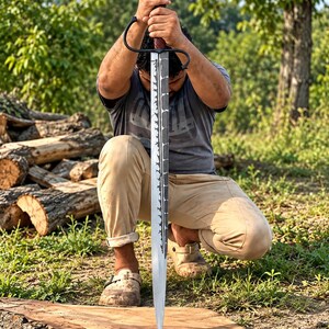 May include: A longsword with a dark, textured blade featuring a jagged, saw-like edge pattern along the center. The sword has a black crossguard, a dark brown spiraled grip, and a rounded pommel, held vertically with the tip resting on wood.