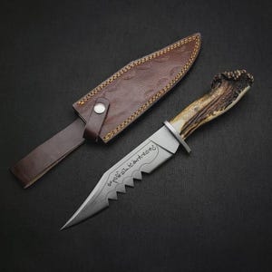 May include: A hunting knife with a serrated blade and a handle made from an antler. The knife is accompanied by a brown leather sheath. The blade has an inscription. The knife is on a black surface.