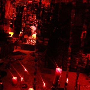 May include: A dimly lit room with a red hue, featuring lit candles and decorative elements. The scene evokes a mystical or spiritual atmosphere, with a focus on the warm glow of the candles.