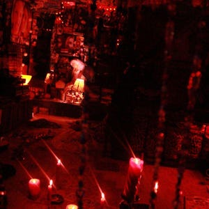 May include: A dimly lit room with a red hue, featuring lit candles and decorative elements. The scene evokes a mystical or spiritual atmosphere, with a focus on the warm glow of the candles.