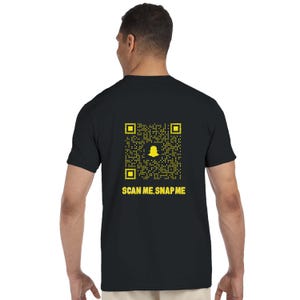 Op de afbeelding: Zwart T-shirt met een gele Snapchat QR-code en de tekst "SCAN ME, SNAP ME". Het ontwerp bevat een geel Snapchat spooklogo in de QR-code. Het shirt heeft korte mouwen en een ronde hals.