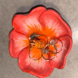 Pode incluir: Um porta-anéis artesanal em forma de flor em tons de vermelho, laranja e branco. O prato contém vários anéis de arame dourado, cada um adornado com pequenas pedras preciosas. O design geral é artístico e decorativo.