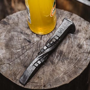 May include: A handcrafted, dark metal bottle opener with a twisted design, resting on a wooden surface next to a yellow beverage bottle. The opener features a textured grip and a unique, rustic aesthetic.