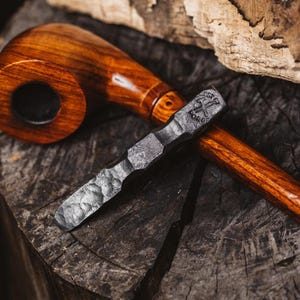 May include: A wooden pipe and a metal tamper rest on a dark wooden surface. The pipe is a rich brown color with a curved stem. The tamper is a dark gray color and has the text "Forge" on it.