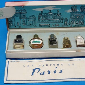 May include: A vintage perfume set with five miniature bottles in a white box, decorated with a blue Parisian cityscape. The set includes bottles labelled "Le Dix", "Revillon", "Bandit", and "Lucien Lelong". A fabric label reads "Les Parfums de Paris".