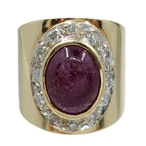 Puede incluir: Un anillo de oro con una gran piedra preciosa ovalada de color rojo oscuro en el centro, rodeada por un halo de pequeñas piedras preciosas claras. El anillo tiene una banda ancha y lisa.