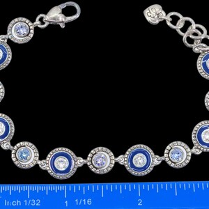 May include: Silver-toned bracelet featuring alternating blue enamel and crystal-studded circular links. The bracelet has a lobster clasp closure and a decorative heart charm. The design incorporates various shades of blue and clear crystals.