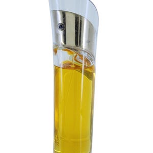 Op de afbeelding: Een heldere glazen parfumfles met een gebogen, zilverkleurige bovenkant en een gouden vloeistof binnenin. De fles heeft een uniek, asymmetrisch ontwerp en een heldere glazen basis. De parfumfles staat rechtop tegen een witte achtergrond.