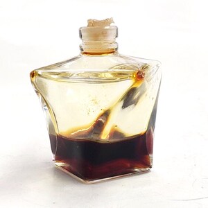 May include: A small, clear glass bottle with a unique, angular shape. The bottle contains a dark brown liquid, with a lighter amber liquid above it. A cork stopper seals the top. The bottle is set against a white background.