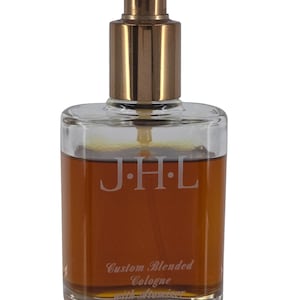 May include: A rectangular glass cologne bottle with a gold-colored sprayer. The bottle contains a brown liquid and has the initials "J.H.L." etched on the front. Below the initials, the text reads "Custom Blended Cologne".