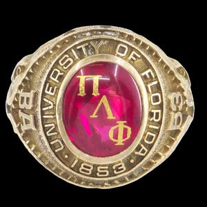 Könnte beinhalten: Ein goldfarbener Abschlussring der University of Florida mit einem rubinroten Mittelstein. Der Ring zeigt die griechischen Buchstaben Π, Λ und Φ sowie das Jahr 1853. Die Worte "UNIVERSITY OF FLORIDA" sind eingraviert.